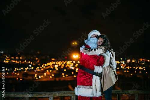 santa claus hugging smiling woman outdoors with lots of christmas lights and christmas presents