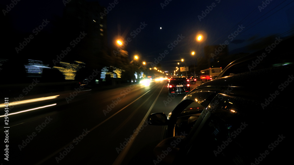 A dark-colored car is moving rapidly along the illuminated street of a night city, on a blurred abstract background a part of the car with a mirror is visible A dark-colored car is moving rapidly along the illuminated street of a night city, on a blurred abstract background a part of the car with a mirror is visible