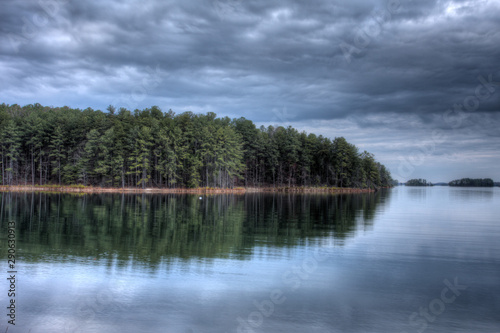 Evergreen trees reflected in the calm waters of a lake with storm clouds overhead as evening approaches.