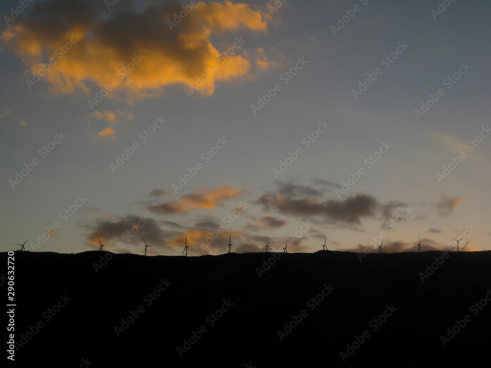 Silhouette of renewable energy wind turbines along ridgeline with sun setting over horizon lighting clouds