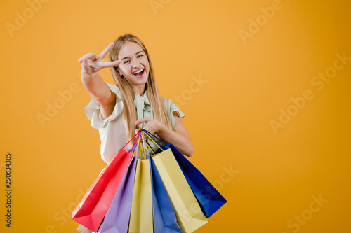happy smiling pretty woman showing peace gesture with colorful shopping bags isolated over yellow