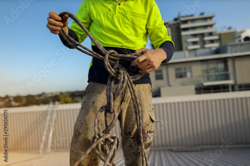Closeup pic of rope access abseiler construction worker standing on top of the roof conducting safety inspecting uncoiling twisting rope prior used construction site Sydney, Australia