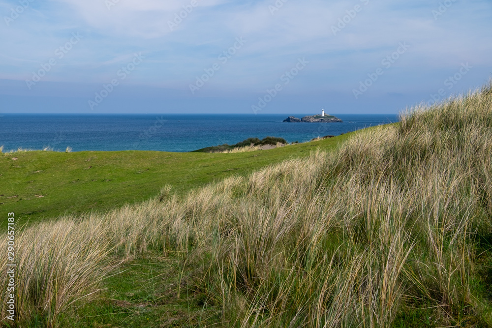 Long grass grows old and yellow as it overlooks a white lighthouse on an island in the middle of the sea, on this partially cloudy day in England, UK.