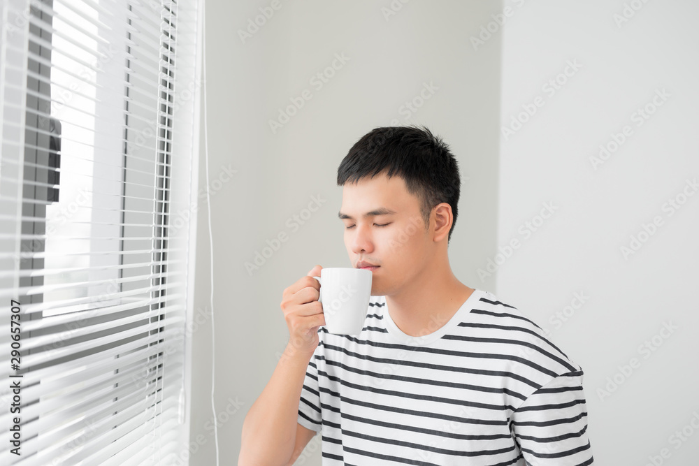 Young handsome carefree man near modern full length window enjoying a cup of coffee while looking outside, good morning, Young handsome carefree man near modern full length window enjoying a cup of coffee while looking outside, good morning,