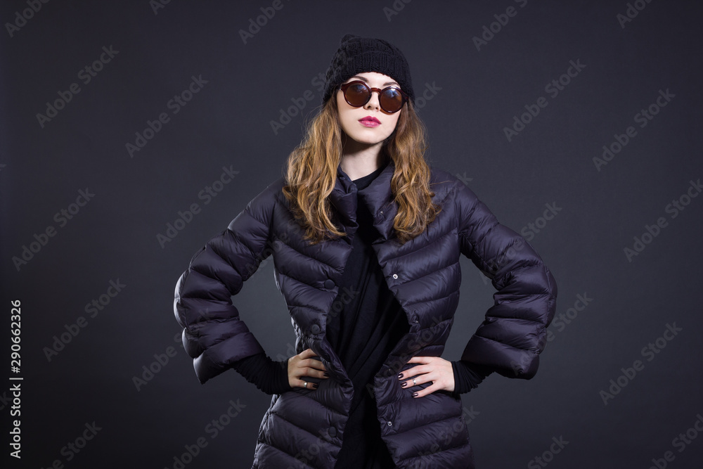 A stylish young woman in a black down coat and knitted hat poses on a black background. Autumn and winter women's fashion. A stylish young woman in a black down coat and knitted hat poses on a black background. Autumn and winter women's fashion.