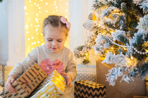 A little beautiful girl sits and opens gifts under a decorated Christmas tree.