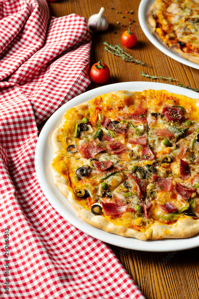 Fresh homemade pizza with prosciutto, capers, olives on a wooden background in composition with a red cloth and olive oil. Italian Cuisine. Selective focus on center of pizza Fresh homemade pizza with prosciutto, capers, olives on a wooden background in composition with a red cloth and olive oil. Italian Cuisine. Selective focus on center of pizza