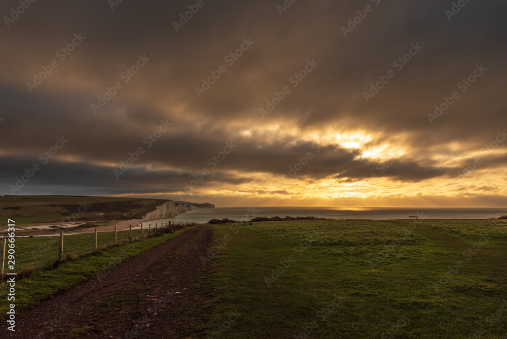 A fence with steel mesh and wooden posts with a dirt road adjacent both go into the horizon as rising sun lights up the clouds and white chalk cliffs famously called the Seven Sisters in England, UK. A fence with steel mesh and wooden posts with a dirt road adjacent both go into the horizon as rising sun lights up the clouds and white chalk cliffs famously called the Seven Sisters in England, UK.