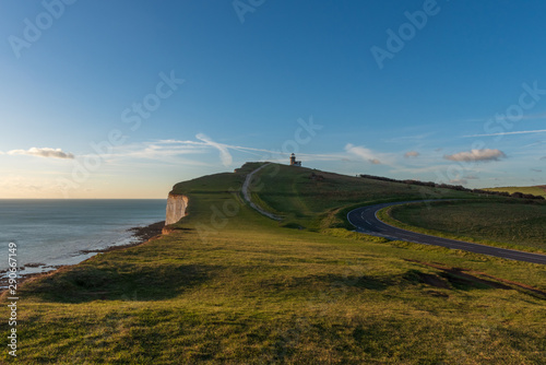 Grassy cliffs with a road that curves around the green fields and footpath that leads to Belle Tout Lighthouse which overlooks the sea on this sunny day with spots of clouds in Eastbourne, UK.