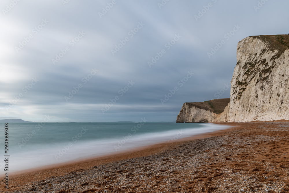 Long exposure of waves crashing into the beach and white chalk cliffs on a cloudy day in England, UK.