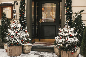 Christmas street decor. Stylish christmas trees with red and gold baubles and festive lights, covered with snow, at front of store or shop at holiday market in city street. Space for text