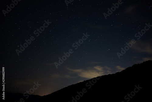 Night starry sky over the mountains. Bright stars and constellations are visible through light clouds.