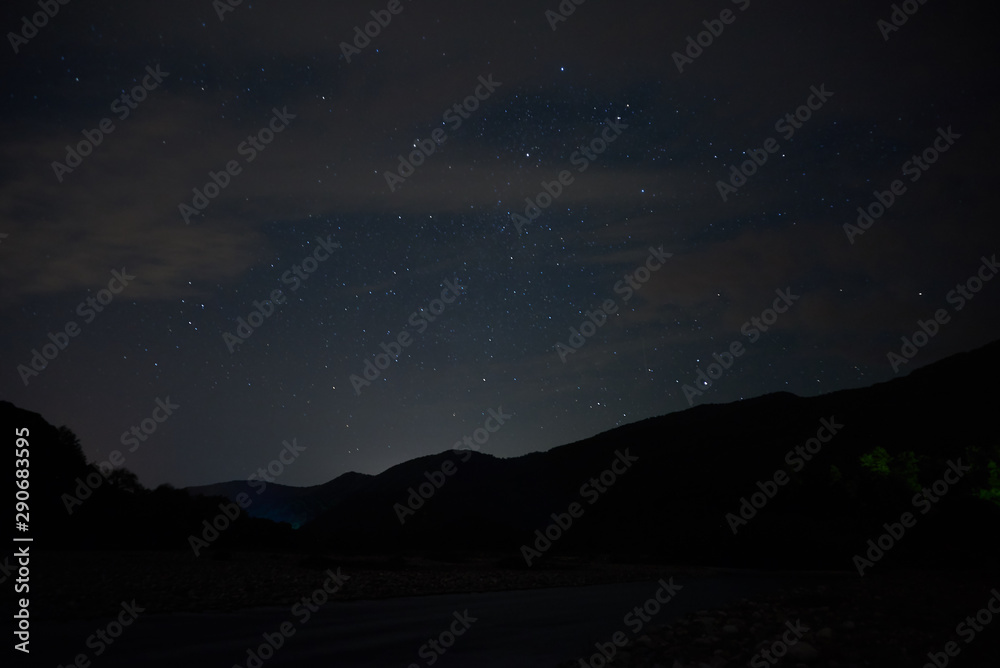 Night starry sky over the mountains. Bright stars and constellations are visible through light clouds.