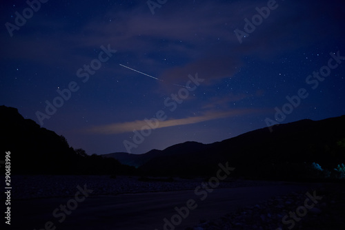 Falling stars. A meteor flies in the night starry sky over the valley of a mountain river.