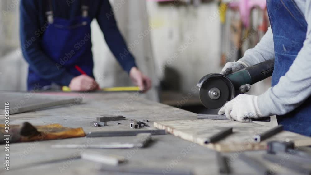 Tracking shot of unrecognizable metalworker cutting metal with angle grinder, his defocused coworker measuring metal plank with measure tape and making marks with pencil in background Tracking shot of unrecognizable metalworker cutting metal with angle grinder, his defocused coworker measuring metal plank with measure tape and making marks with pencil in background