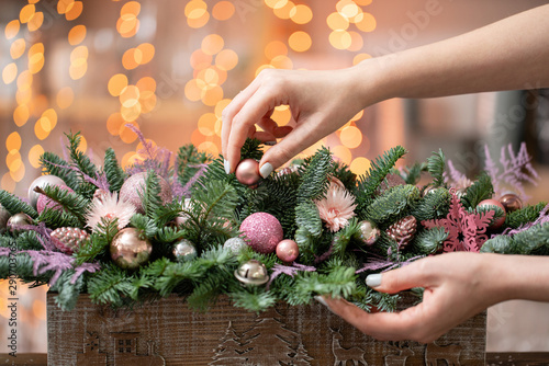 Young woman creates and decorates beautiful festive arrangement of fresh spruce  ornamentals in a rustic wooden box box. Christmas mood. Garland bokeh on background.