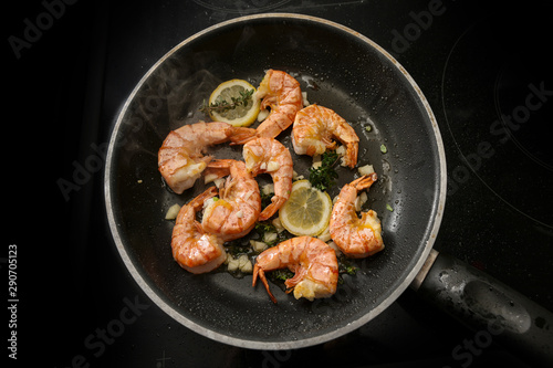 tiger prawn shrimps are roasted in a black pan with garlic, lemon and herbs on a black stove, high angle view from above