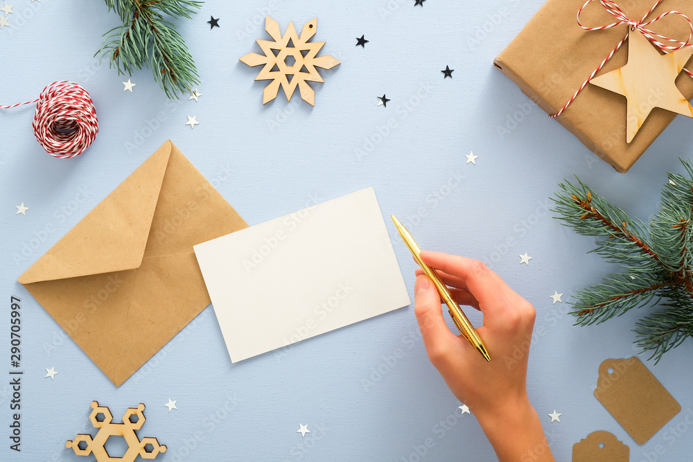 Female hand writing letter to Santa Claus over blue background with festive Christmas decorations, confetti, fir tree branches, snowflakes, gift box. Christmas, New Year, winter holiday concept Female hand writing letter to Santa Claus over blue background with festive Christmas decorations, confetti, fir tree branches, snowflakes, gift box. Christmas, New Year, winter holiday concept