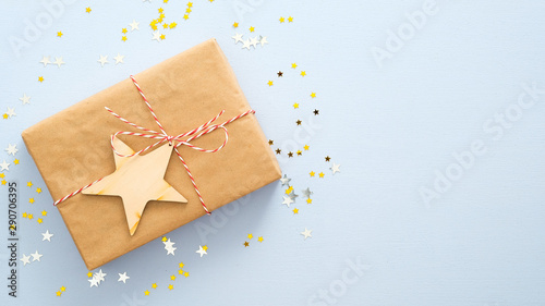Flat lay gift box with striped red ribbon and wooden star over pastel blue background with golden confetti stars. Christmas, New Year, winter holidays and birthday concept. Top view with copy space