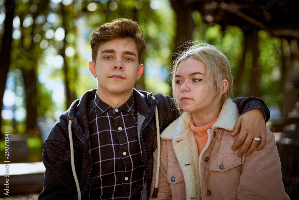 Teenagers in love sit on park bench, resting in rays of autumn sun, boy looking straight ahead. Concept of teen love