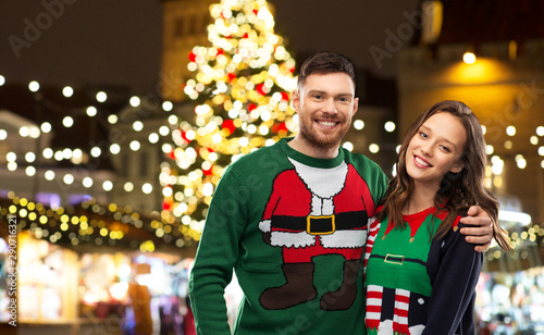 winter holidays, celebration and people concept - portrait of happy couple in ugly sweaters hugging over christmas market lights background