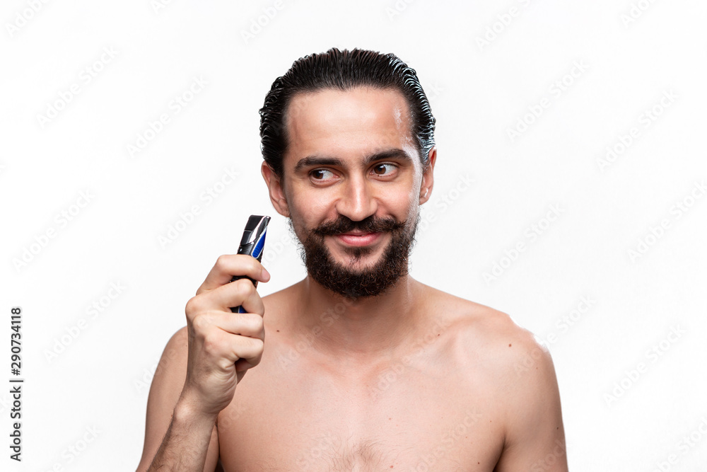 Excited bearded man with mustache holds electric shaver standning bare isolated over white background. Concept of morning treatment and shaving. Time to trim your beard. Morning routine