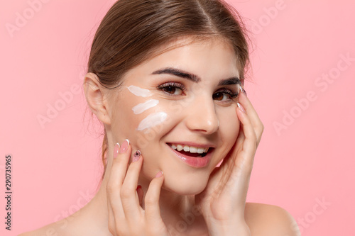 Beautiful model applyes moisturizing cosmetic product to skin holding white tube of a hyaluronic cream isolated over pink background. Concept of beauty and health treatment.