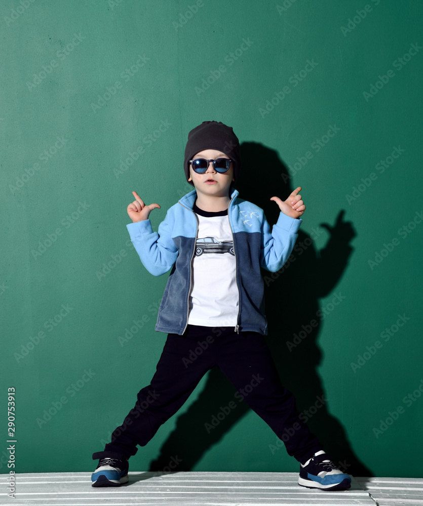Cool boy in blue sunglasses, headwear, fleece jacket, pants and sneakers is posing like a gangster with two finger guns