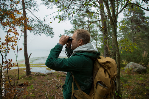Mature man exploring Finland in the fall, looking into fog through binoculars. Hiker with big backpack standing on mossy rock. Scandinavian landscape with misty sea and autumn forest. © Suzi Media 