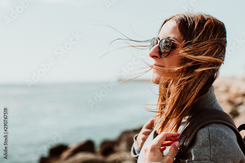 .Pretty young woman with grey coat enjoying a sunny and windy morning around the coast in Porto, Portugal. Walking relaxed, happy and carefree. Lifestyle photo
