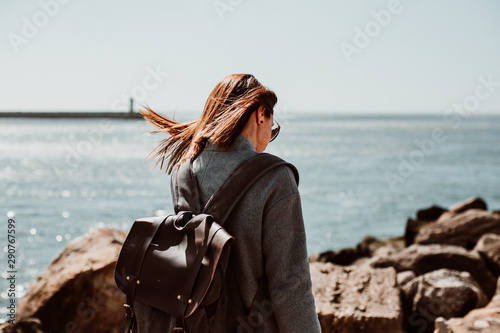 .Pretty young woman with grey coat enjoying a sunny and windy morning around the coast in Porto, Portugal. Walking relaxed, happy and carefree. Lifestyle