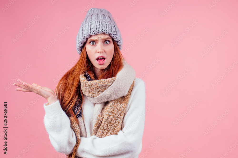 amazed redhaired ginger woman wear stylish wool hat ,sweater and scarf looking at camera and gesturing pointing fingers at copyspase in studio pink background