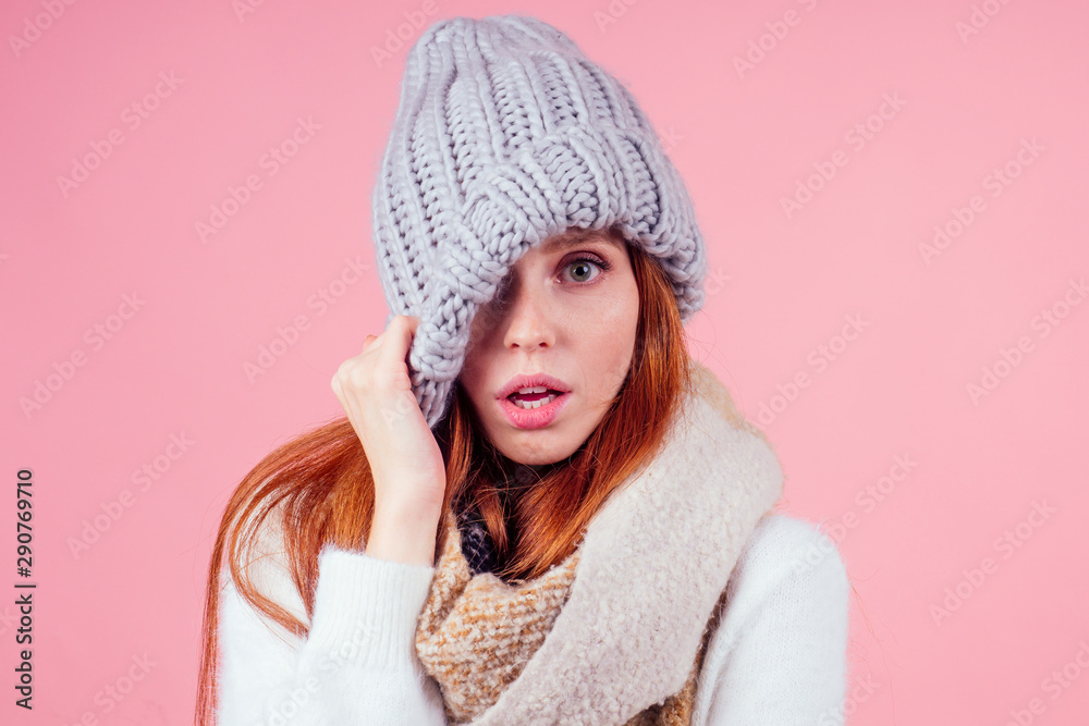 shy redhead ginger woman putting a knitted spiky hat on his head,hiding eye and open mouth in studio pink background shy redhead ginger woman putting a knitted spiky hat on his head,hiding eye and open mouth in studio pink background