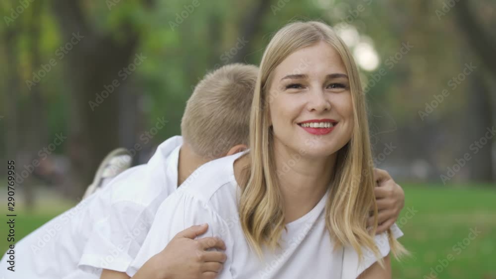 Portrait of attractive caucasian woman in the park looking at camera and smiling. Two little children running towards her from the background and hugging their mother. Sunny day, leisure outdoors. Portrait of attractive caucasian woman in the park looking at camera and smiling. Two little children running towards her from the background and hugging their mother. Sunny day, leisure outdoors.