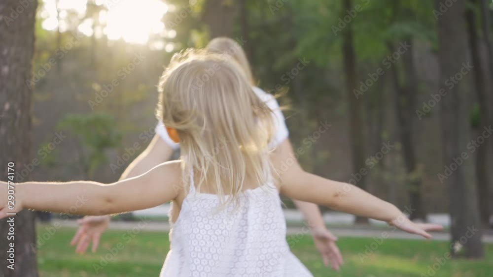 Back view of little blond girl running towards her mother in the sunshine. Attractive young woman hugging her daughter and smiling. Happy family spending time together in the park. Slow motion. Back view of little blond girl running towards her mother in the sunshine. Attractive young woman hugging her daughter and smiling. Happy family spending time together in the park. Slow motion.