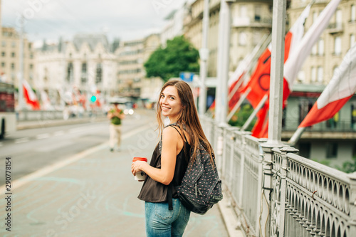 Outdoor portrait of beautiful young woman in the city, wearing backpack, holding cup of take away coffee. Image taken in Lausanne downtown, place Bel Air, Switzerland