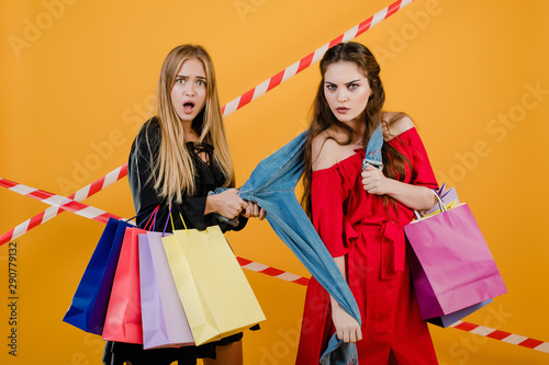 pretty girls fighting over pair of jeans with colorful shopping bags and signal tape isolated over yellow