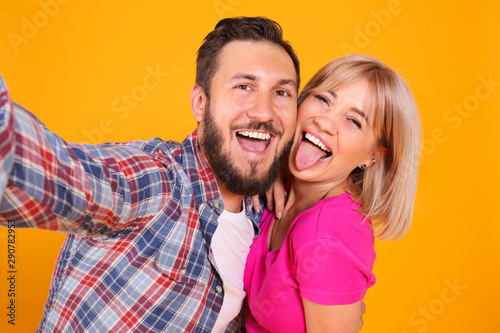 Young beautiful hipster couple having fun posing over isolated yellow background. Portrait of tall man with groomed beard and his short attractive blonde girlfriend. Copy space, close up.