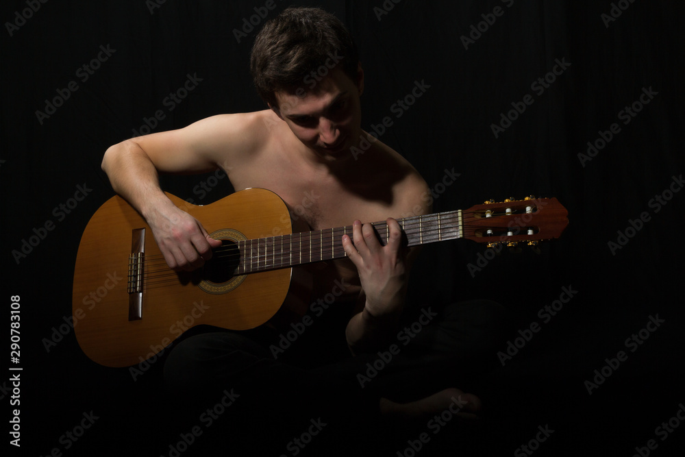Obraz premium Handsome young musician playing on guitar. Naked torso. On black background.
