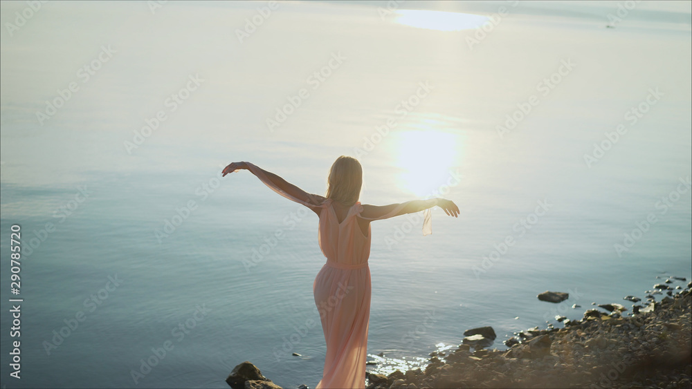 Beautiful young woman standing in sea with waves on sandy beach against orange sky at sunset. Summer travel. A young girl is standing against the sunset in a summer dress. Beautiful young woman standing in sea with waves on sandy beach against orange sky at sunset. Summer travel. A young girl is standing against the sunset in a summer dress.