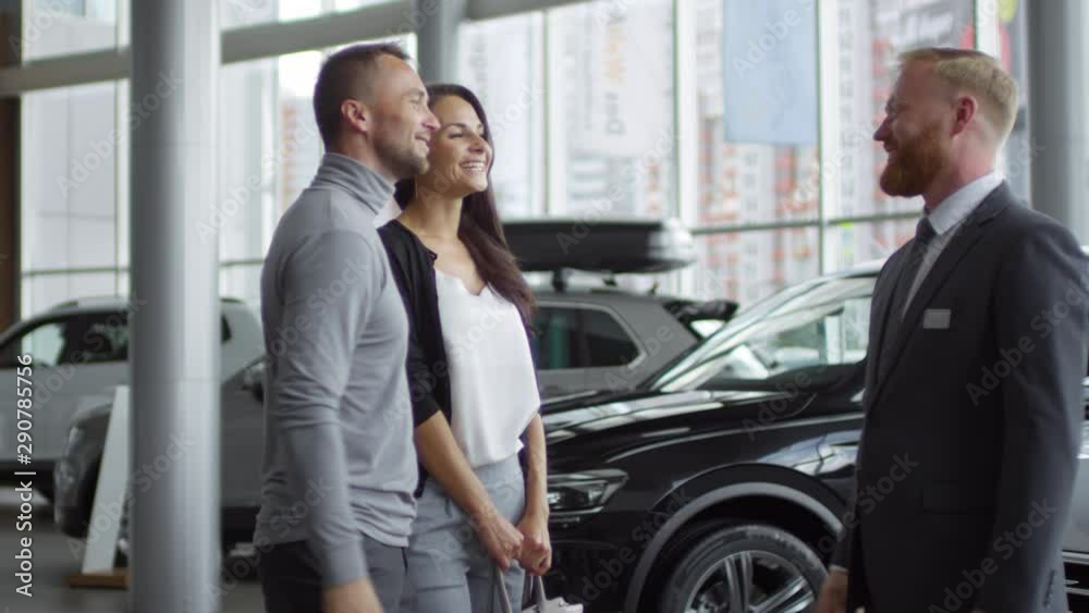 Cheerful seller shaking hands with male customer and talking with his smiling wife after making successful deal in car showroom