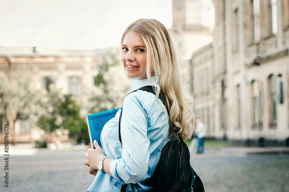 A blonde student girl is smiling and holding a folder and a notebook in her hands at university background. Girl is taking exams at university