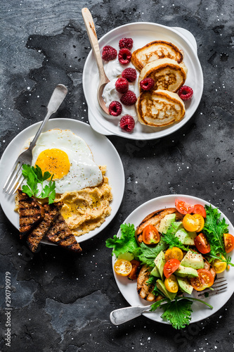 Savory and sweet breakfast - avocado, cherry tomatoes grilled  bread salad, fried egg with hummus and rye croutons and pancakes with greek yogurt and raspberries on dark background, top view