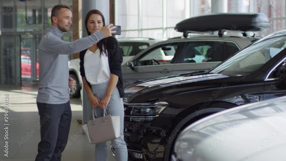 Man taking picture of new car with smartphone and discussing it with beautiful wife while choosing new car in auto dealership showroom Man taking picture of new car with smartphone and discussing it with beautiful wife while choosing new car in auto dealership showroom
