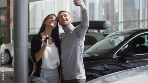Cheerful caucasian couple standing beside new car in auto dealership showroom, smiling and showing key at camera while taking selfie with smartphone