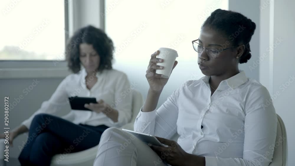Serious businesswomen with digital tablets. Side view of focused multiethnic businesswomen drinking from paper cups and using tablet computers in office. Technology concept Serious businesswomen with digital tablets. Side view of focused multiethnic businesswomen drinking from paper cups and using tablet computers in office. Technology concept