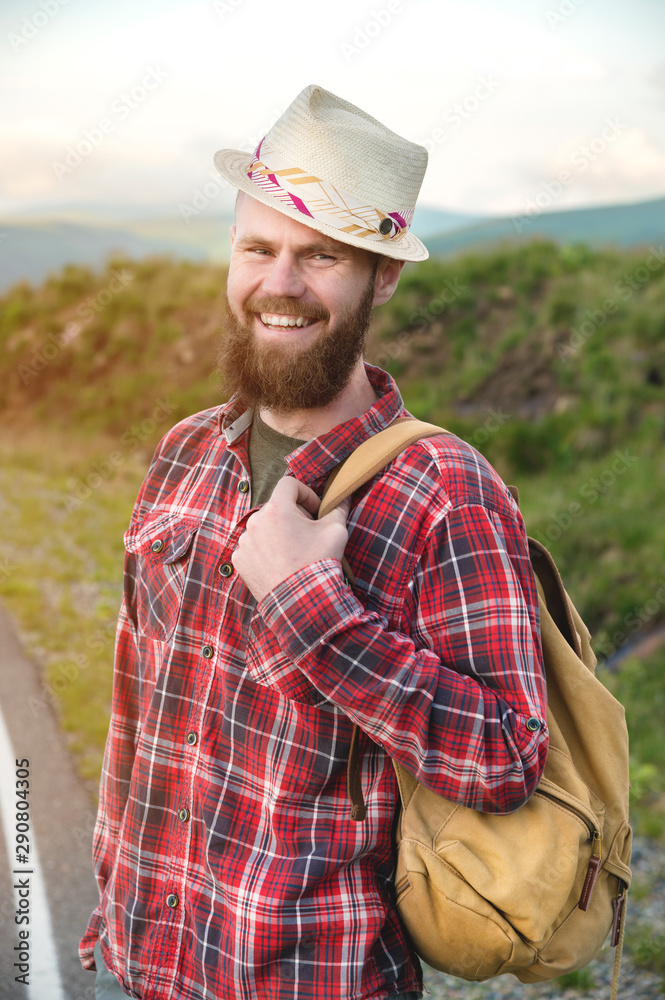Portrait of a bearded happy smiling traveler hipster with a backpack in a plaid shirt and a hat next to an unknown car stands on the road at sunset in the mountains. Happy and confident travel concept Portrait of a bearded happy smiling traveler hipster with a backpack in a plaid shirt and a hat next to an unknown car stands on the road at sunset in the mountains. Happy and confident travel concept