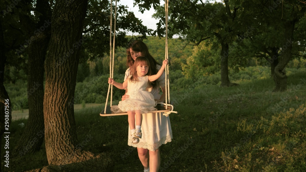 Mom shakes her daughter on swing under a tree in sun. close-up. mother and baby ride on rope swing on an oak branch in forest. Girl laughs, rejoices. Family fun in park, in nature. warm summer day.