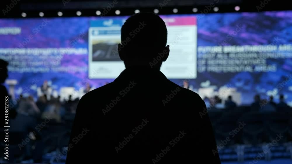 Male speaker goes on stage. Forum on business development. Hall is full. View from the back of auditorium with people at a conference or presentation. International economic business forum. Indoors