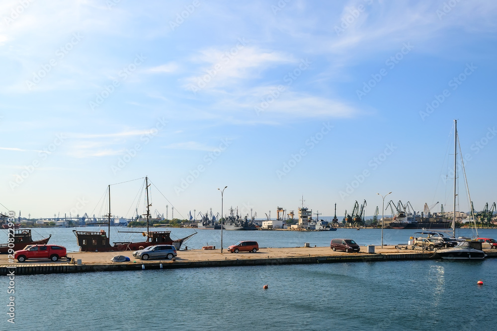 View of the water area of the small seaport with yachts, cargo ships and cranes on a clear and calm sunny day. Industrial seascape.